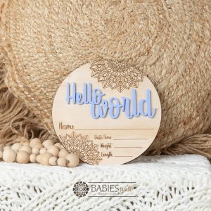 Round timber Birth Announcement Plaque featuring purple raised acrylic "Hello World" lettering and engraved mandela floral designs, with spaces to record a baby's birth detals. Styled on a soft white blanket with a woven pillow & wooden beads.