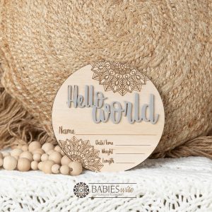 Round timber Birth Announcement Plaque featuring Mocha raised acrylic "Hello World" lettering and engraved mandela floral designs, with spaces to record a baby's birth detals. Styled on a soft white blanket with a woven pillow & wooden beads.