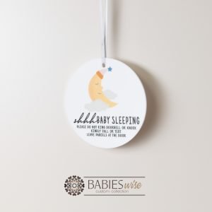 Round white acrylic plaque with hole for hanging on a door, UV printed with "shhh... baby sleeping with message and a sleeping half moon illusration. Styled hanging by a ribbon.