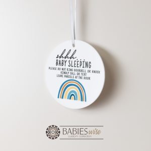 Round white acrylic plaque with hole for hanging on a door, UV printed with "shhh... baby sleeping with message and a blue toned rainbow. Styled hanging by a ribbon.