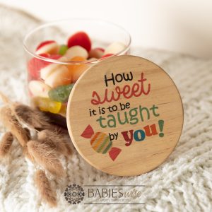 Glass gift jar with UV printed timber lid reading "How sweet it is to be taught by you". Styled with lollies in the jar on a soft blanket.