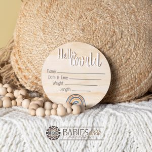 Round timber "Hello World" birth announcement plaque with UV printed text and rainbow design featuring spaces for baby's name, date, time, weight and lenght. Styled with a soft blanket and woven pillow.