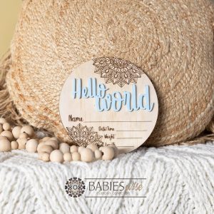 Round timber Birth Announcement Plaque featuring blue raised acrylic "Hello World" lettering and engraved mandela floral designs, with spaces to record a baby's birth detals. Styled on a soft white blanket with a woven pillow & wooden beads.