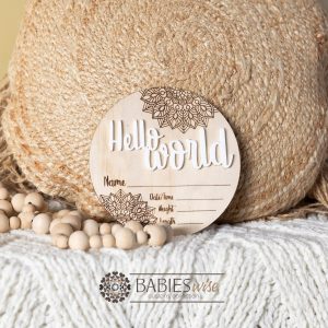 Round timber Birth Announcement Plaque featuring white raised acrylic "Hello World" lettering and engraved mandela floral designs, with spaces to record a baby's birth detals. Styled on a soft white blanket with a woven pillow & wooden beads.