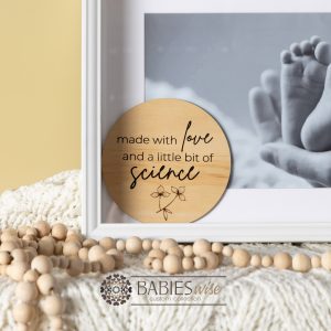 Round timber plaque engraved with the words "made with love and a little bit of science" in mixed fonts with a small floral line drawing at the bottom. Styled with a soft blanket and set in a frame with the baby's feet.
