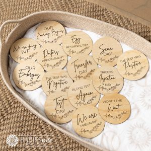 IVF pregnancy milestone plaques UV printed on timber with delicate leaf motif, showing fertility journey stages from IVF to pregnancy, Styled on a woven bassinett on a woven neutral mat.