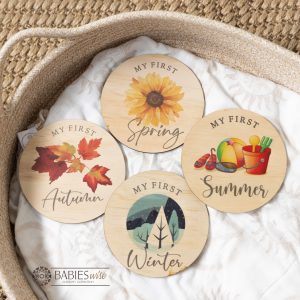 Set of four round timber milestone plaques UV Printed with "MY FIRST, Summer, Autumn, Winter, Spring". Featuring mixed script and bright illustration on each plaque. Styled on a soft blanket in a woven basket.