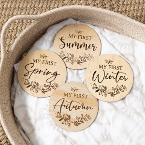 Set of four round timber milestone plaques engraved with "MY FIRST, Summer, Autumn, Winter, Spring". Featuring mixed script and florals with a bird. Styled on a soft blanket in a woven basket.