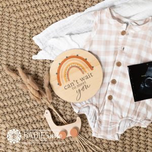 Round timber pregnancy announcement plaque with brown rainbow design and "can't wait to meet you" text. Styled on a woven mat with baby outfit and ultrasound photo.