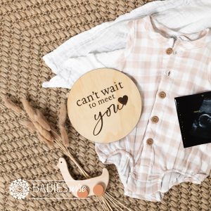 Round timber pregnancy announcement plaque laser engraved with "can't wait to meet you" text and small loveheart. Styled on a woven mat with a baby outfit and ultrasound photo.
