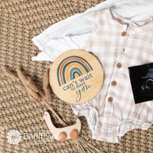 Round timber pregnancy announcement plaque with blue rainbow design and "can't wait to meet you" text. Styled on a woven mat with baby outfit and ultrasound photo.