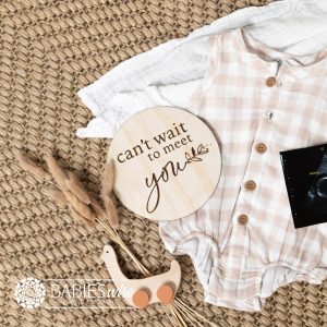 Round timber pregnancy announcement plaque laser engraved with "can't wait to meet you" text and small loveheart. Styled on a woven mat with a baby outfit and ultrasound photo.