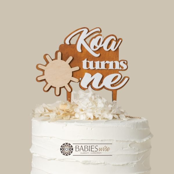 Personlised first birthday cake topper featuring a timber sun design, a custom name, and raised 3D acrylic lettering reading "turns one". Styled on a white cake with flowers.