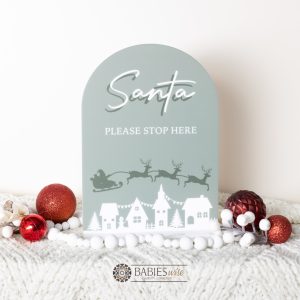 Olive acrylic Christmas plaque reading Santa Please Stop Here with sleigh and reindeer flying over village scene. Styled iwith a soft blanket and Christmas decorations.