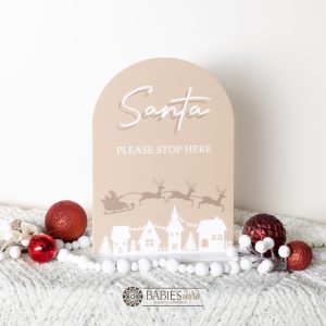 Latte acrylic Christmas plaque reading Santa Please Stop Here with sleigh and reindeer flying over village scene. Styled iwith a soft blanket and Christmas decorations.