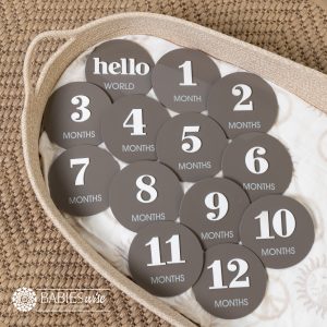 Baby monthly milestone discs in coffee acrylic with raised white 3D numbers and printed months text. Beginning with "Hello World, 1 month to 12 months. Styled on a woven bassinett on a woven neutral mat.