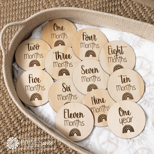 Engraved timber baby milestone discs from one month to one year, each featuring a small rainbow design and script lettering. Displayed on a soft blanket in a wicker basket.