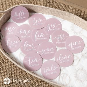 Baby Monthly milestone discs in a dusty soft pink acrylic with white flowing cursive script. Styled on a woven bassinett on a woven neutral mat.