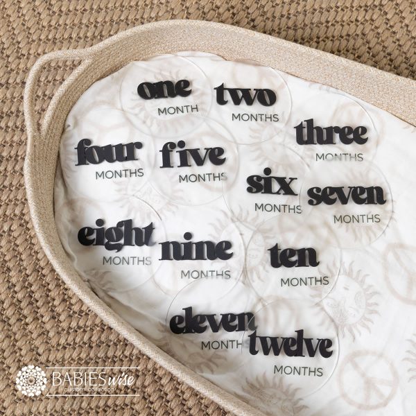 Baby monthly milestone discs in frosted acrylic with raised 3D black bold in lower case modern lettering. Displayed on a soft blanket in a wicker basket.
