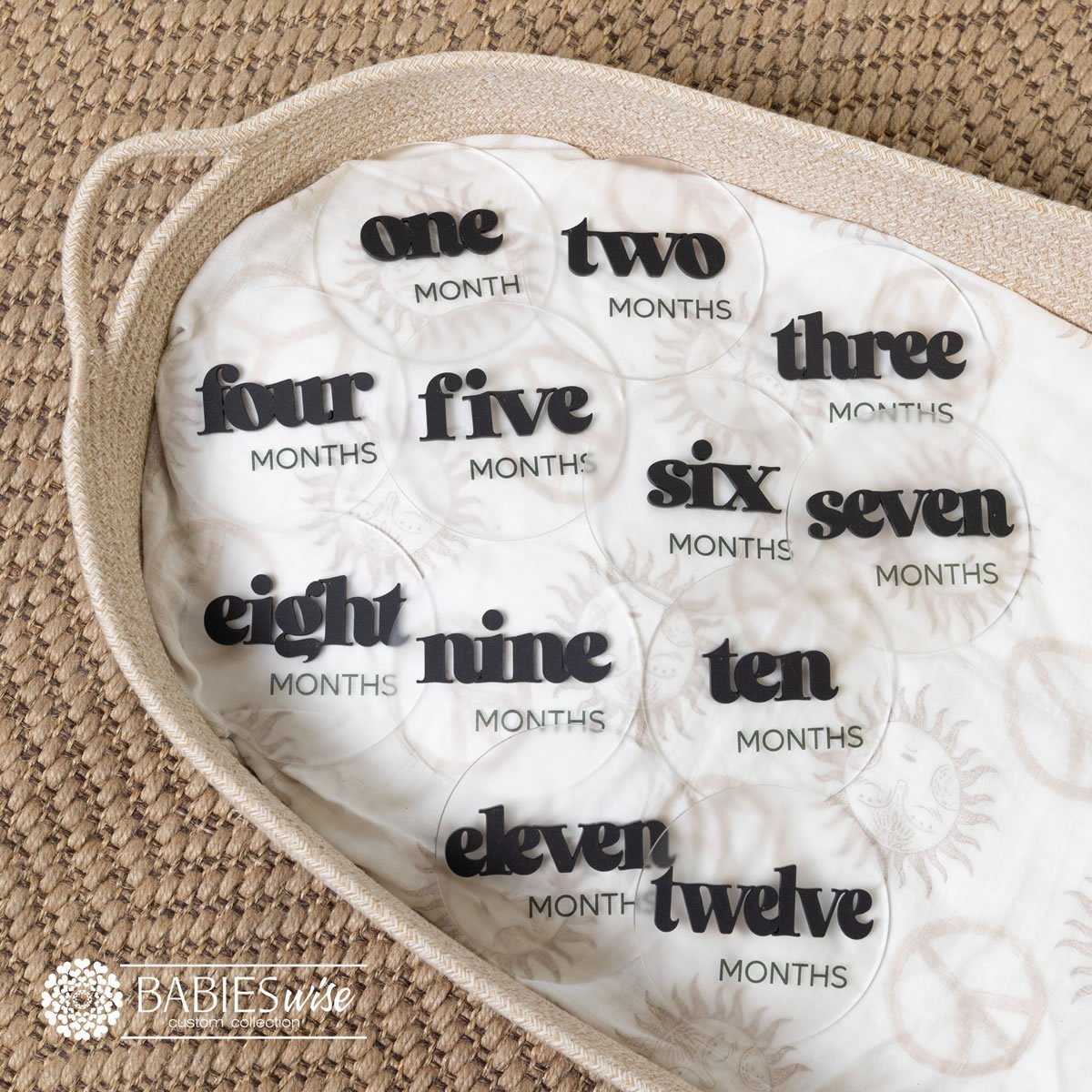 Baby monthly milestone discs in frosted acrylic with raised 3D black bold in lower case modern lettering. Displayed on a soft blanket in a wicker basket.