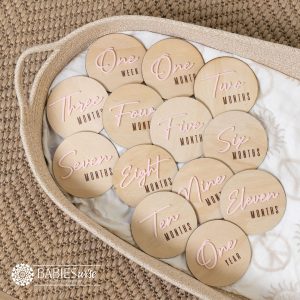 Baby milestone discs with natural timber base and raised baby pink 3D acrylic flowing cursive lettering from one week, months and one year. Styled on a woven bassinett on a woven neutral mat.