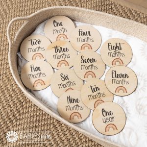 Baby monthly milestone discs UV printed on timber with a Brown rainbow design and black script text. Styled on a woven bassinett on a woven neutral mat.