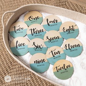 Baby monthly milestone discs with the top half in a natural timber and the bottom half UV printed in a soft teal colour, featuring black script months text. Styled on a woven bassinett on a woven neutral mat.