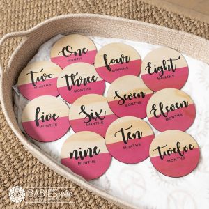 Baby monthly milestone discs with the top half natural timber and the bottom half UV printed in vibrant pink colour featuring black script text. Styled on a woven bassinett on a woven neutral mat.
