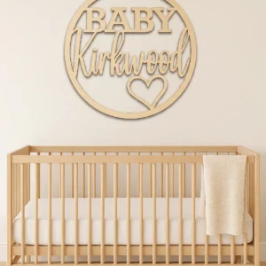 Personlised circular timber nursery sign featyring the word BABY in bold uppercase lettering, the surname is in a script font, and a love heart shape within a timber hoop. Styled in a nursery sign is above the neutral cot.