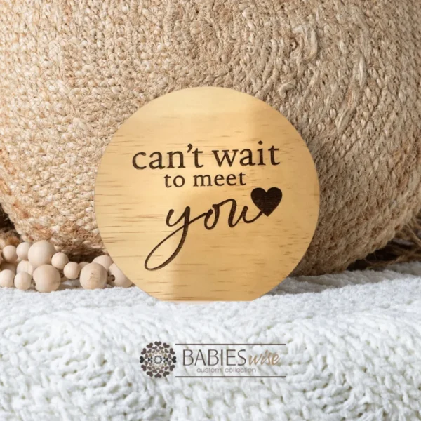 Round timber pregnancy announcement plaque laser engraved with "can't wait to meet you" text and small loveheart. Styled on soft blanket with a woven pillow and wooden beads.