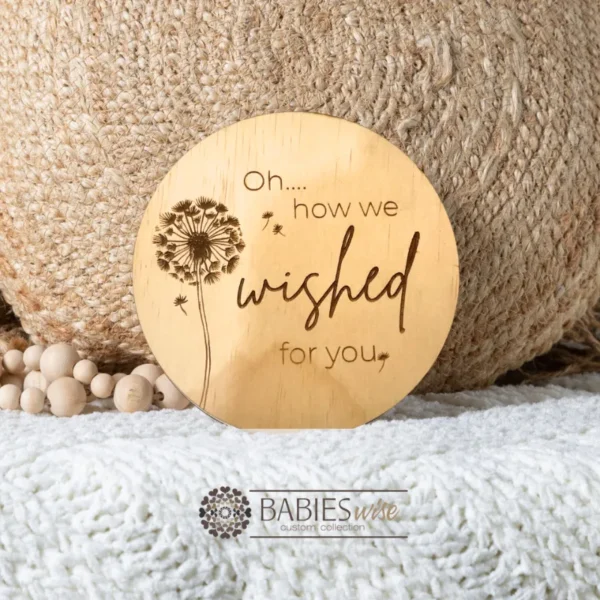 Round timber plaque laser engraved with the words "oh... how we wished for you" with a Dandelion. Style on a soft blanket with a woven pillow and wooden beads.