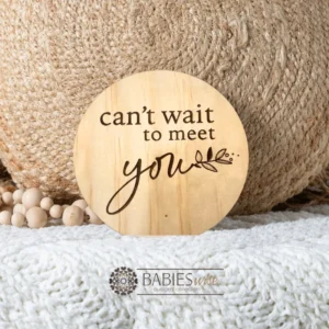 Round timber pregnancy announcement plaque laser engraved with "can't wait to meet you" text and small loveheart. .Styled on soft blanket with a woven pillow and wooden beads.
