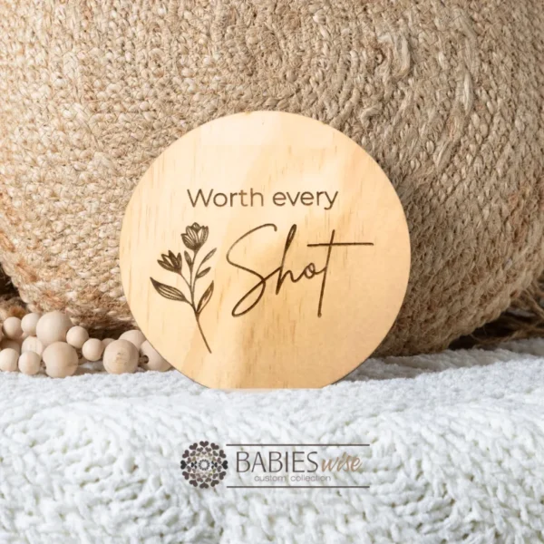 Round timber plaque engraved with the words "worth every shot" and a delicate botanical sprig design. Styled on a soft blanket with a woven pillow and wooden beads.