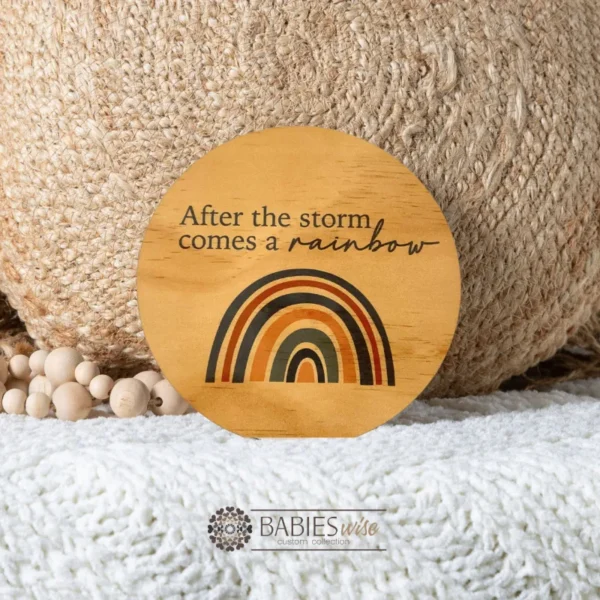 Round timber plaque with UV printed text "After the storm comes a rainbow" and a rainbow illustration in blue and brown tones. Styled on a soft blanket with a woven pillow and wooden beads.
