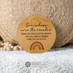 Round timber plaque with printed quote- "somewhere over the rainbow, Skies are blue and the dreams that you dare to dream really do come true". with a small brown-toned rainbow. Styled on a soft blanket withna woven pillow and wooden beads.