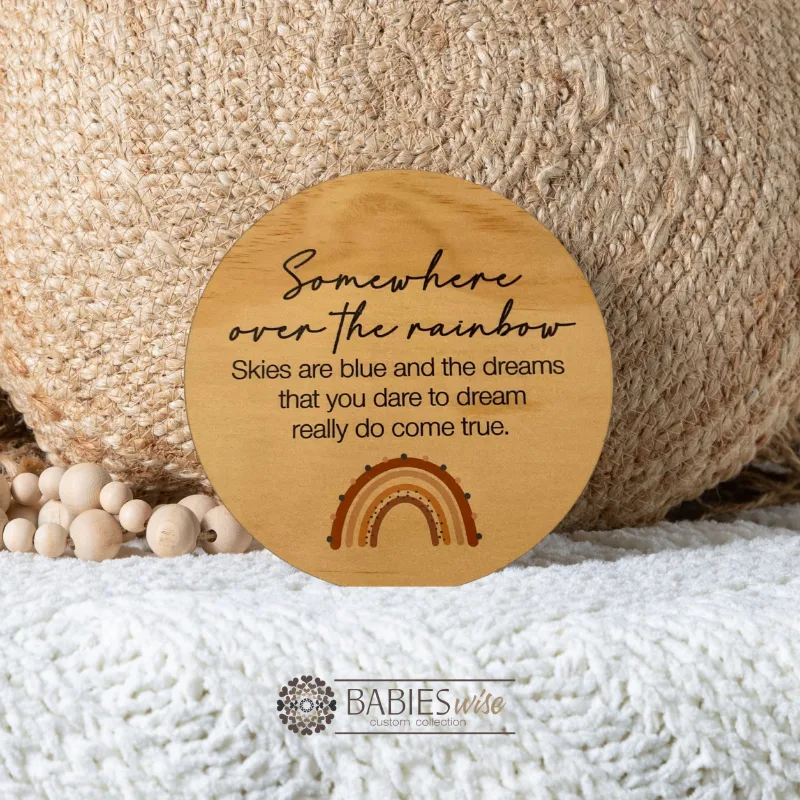 Round timber plaque with printed quote- "somewhere over the rainbow, Skies are blue and the dreams that you dare to dream really do come true". with a small brown-toned rainbow. Styled on a soft blanket withna woven pillow and wooden beads.
