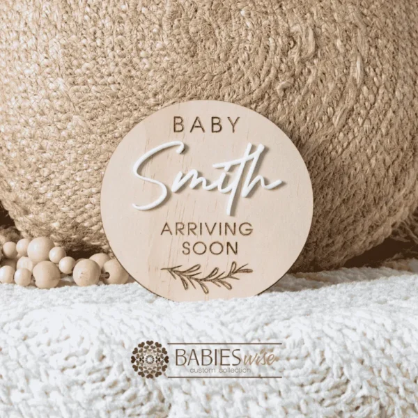 Round timber pregnancy announcement plaque with raised acrylic surname and engraved wording "Baby Arriving Soon" with small leaf illustration. Styled on a soft white blanket with a woven pillow & wooden beads.