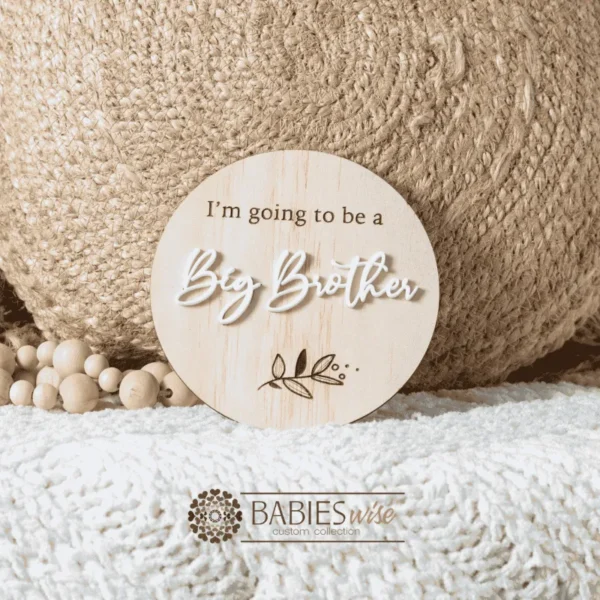 Round natural timber plaque with enraved "I'm going to be a" and raised 3D acryl with lettering reading "Big Brother" and a engraved leaf motif. Styled on a soft white blanket with a woven pillow & wooden beads.