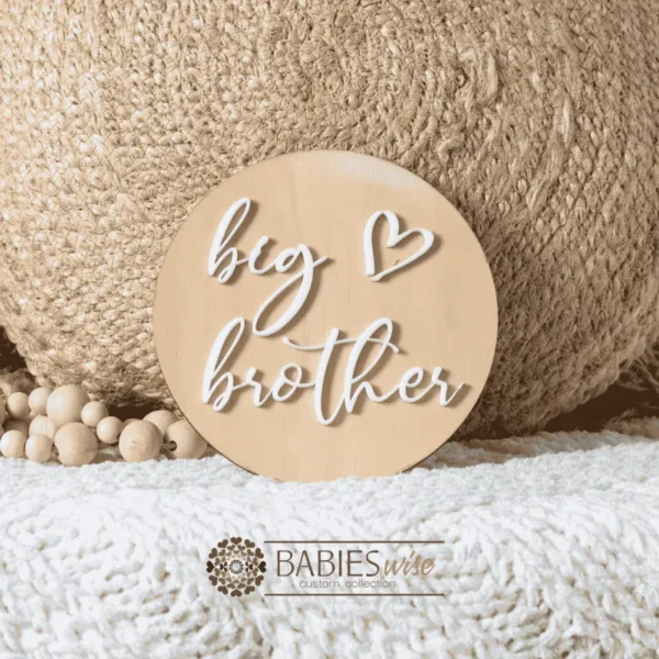 Round timber sibling announcement plaque with raised white acrylic script reading "big brother" and a small loveheart detail. Styled on a soft white blanket with a woven pillow & wooden beads.