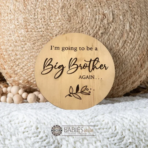 Round laser engraved timber plaque with the words "I'm going to be a Big Brother AGAIN " in moxed script and uppercase lettering with a small botanical leaf design. Styled on a soft white blanket with a woven pillow & wooden beads.