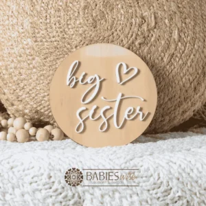 Round timber sibling announcement plaque with raised white acrylic script reading "big brother" and a small loveheart detail. Styled on a soft white blanket with a woven pillow & wooden beads.