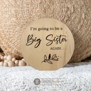 Round laser engraved timber plaque with the words "I'm going to be a Big Sister AGAIN " in moxed script and uppercase lettering with a small botanical leaf design. Styled on a soft white blanket with a woven pillow & wooden beads.