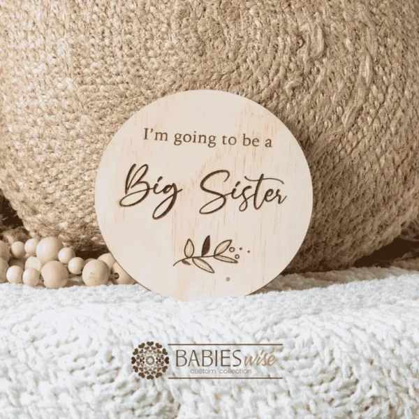 Round laser engraved timber plaque with the words "I'm going to be a Big Sister" in moxed script and uppercase lettering with a small botanical leaf design. Styled on a soft white blanket with a woven pillow & wooden beads.