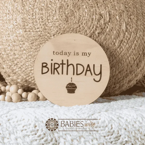 Round laser engraved timber milestone plaque with the words "today is my Birthday" and a small cupcake illustration underneath. Styled on a soft white blanket with a woven pillow & wooden beads.