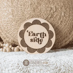 Round natural timber plaque laser engraved with the words "Earthside" in a decorative vintage-style font, surrounded by a repeating rainbow-inspired scallop pattern. Styled on a soft white blanket with a woven pillow & wooden beads.