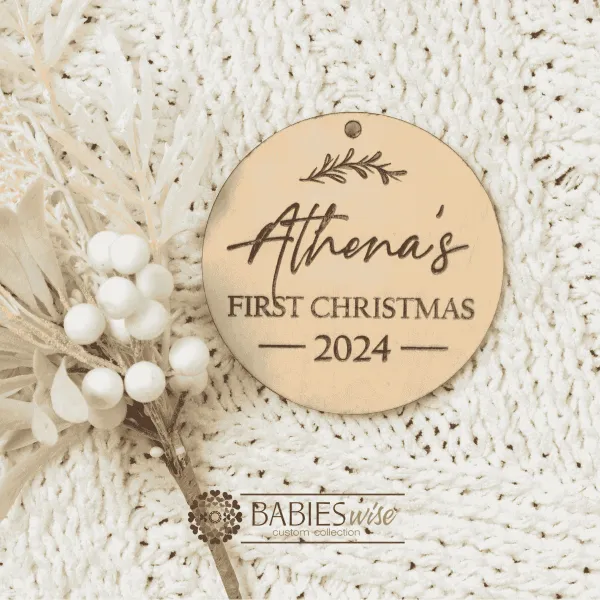 Personlised laser engraved timber First Christmas bauble featuring a child's name, decorative leaf motif leaf and year. Styled on a soft, white blankey with a white wreath.