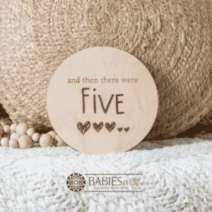 Round natural timber plaque engraved with words "and then there were five" and five small hearts illustration. Styled on a soft white blanket with a woven pillow & wooden beads.