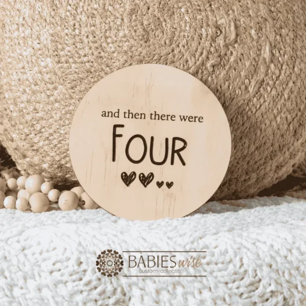 Round natural timber plaque engraved with words "and then there were four" and four small hearts illustration. Styled on a soft white blanket with a woven pillow & wooden beads.