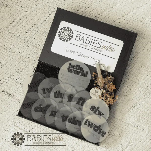 Luxury newborn gift box featuring frosted acrylic monthly milestone discs with black 3D numbers, a matching Hello World announcement plaque, Shh.. Baby Sleeping plaque, and dried flowers in a black magnetic gift box. Styled in a black gift box with black straw on a soft blanket.