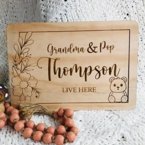 Personlised rectangular timber plaque engraved with grandparents names, surname and the words LIVE HERE. featuring a floral line artwork on the leaf, a cute teddy bear illustration on the right. Styled on a soft white blanket with wooden beads.